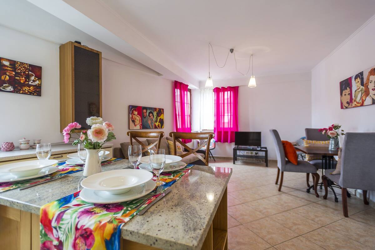 A dining area with a table set for four, chairs, a vase with flowers, and a television in the background.