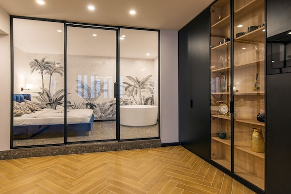 A modern interior with a large sliding glass door leading to a bedroom with a bed and a large bathtub. There is a wooden cabinet with shelves on the right side.