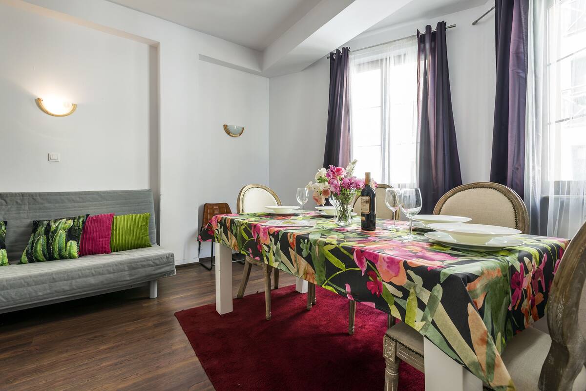 A dining area with a table set for two, a sofa with colorful cushions, and a red rug.
