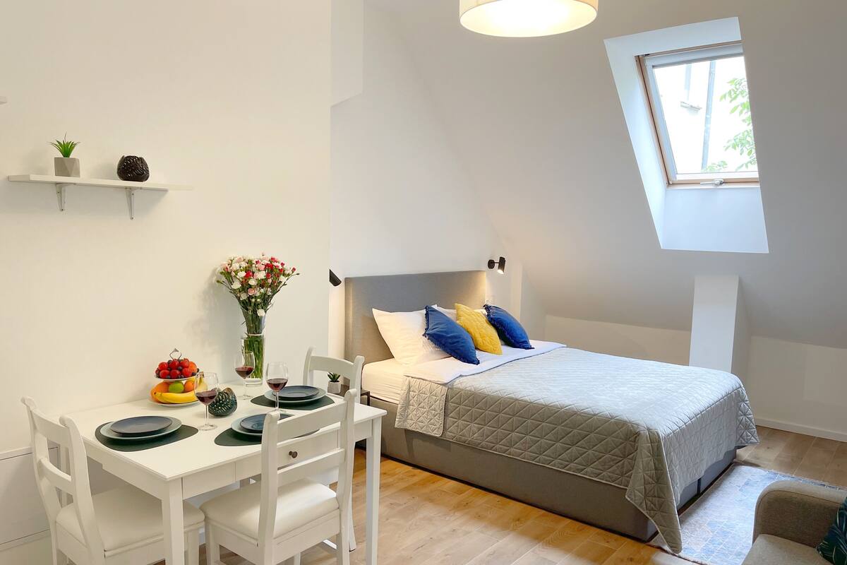 A compact room with a bed, a dining table with chairs, a small shelf with decorative items, and a window with natural light.