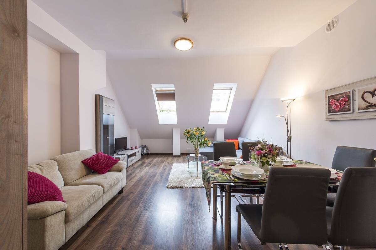 A modern living room with a sofa, dining table, and chairs, a TV, and a ceiling with skylights.