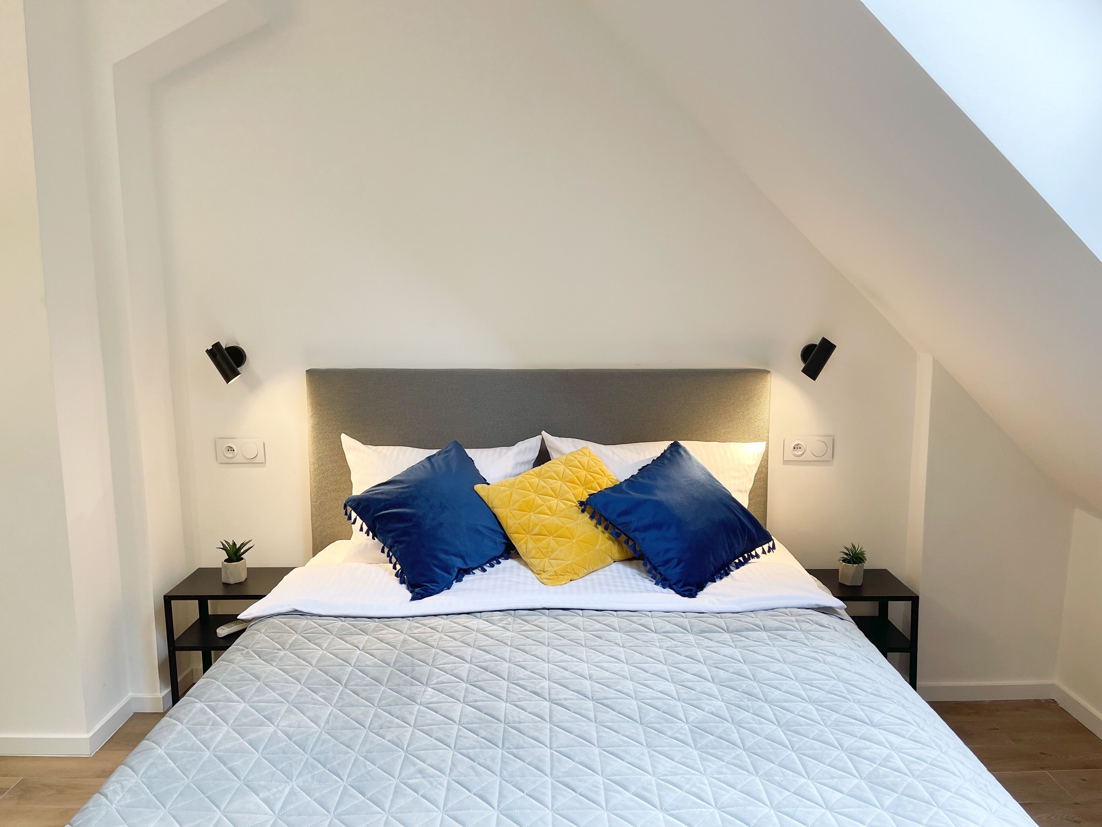 Une chambre à coucher avec un lit, une tête de lit grise et deux tables de chevet ornées de petites plantes.