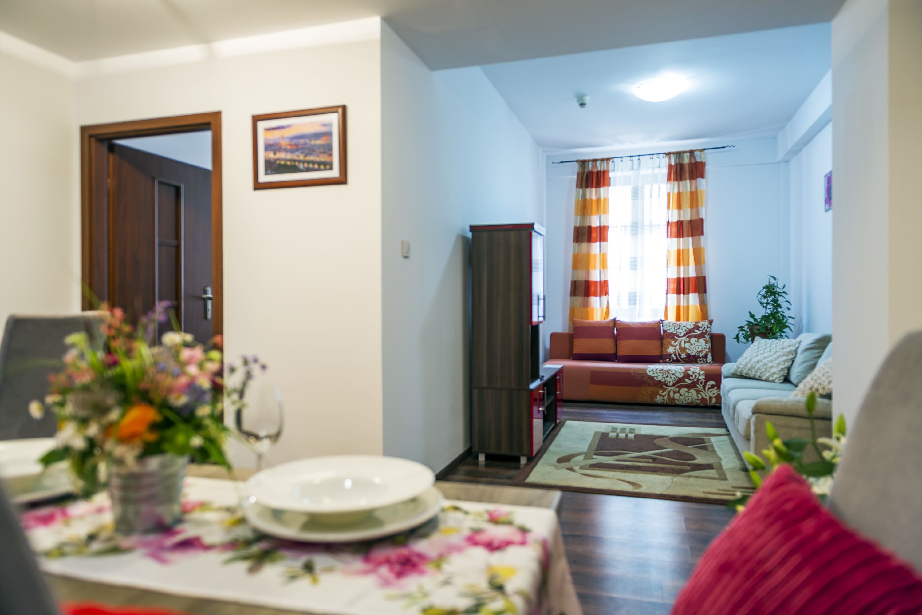 A living room with a sofa, a dining table with plates and a vase of flowers, and a view into the adjacent room through an open door.