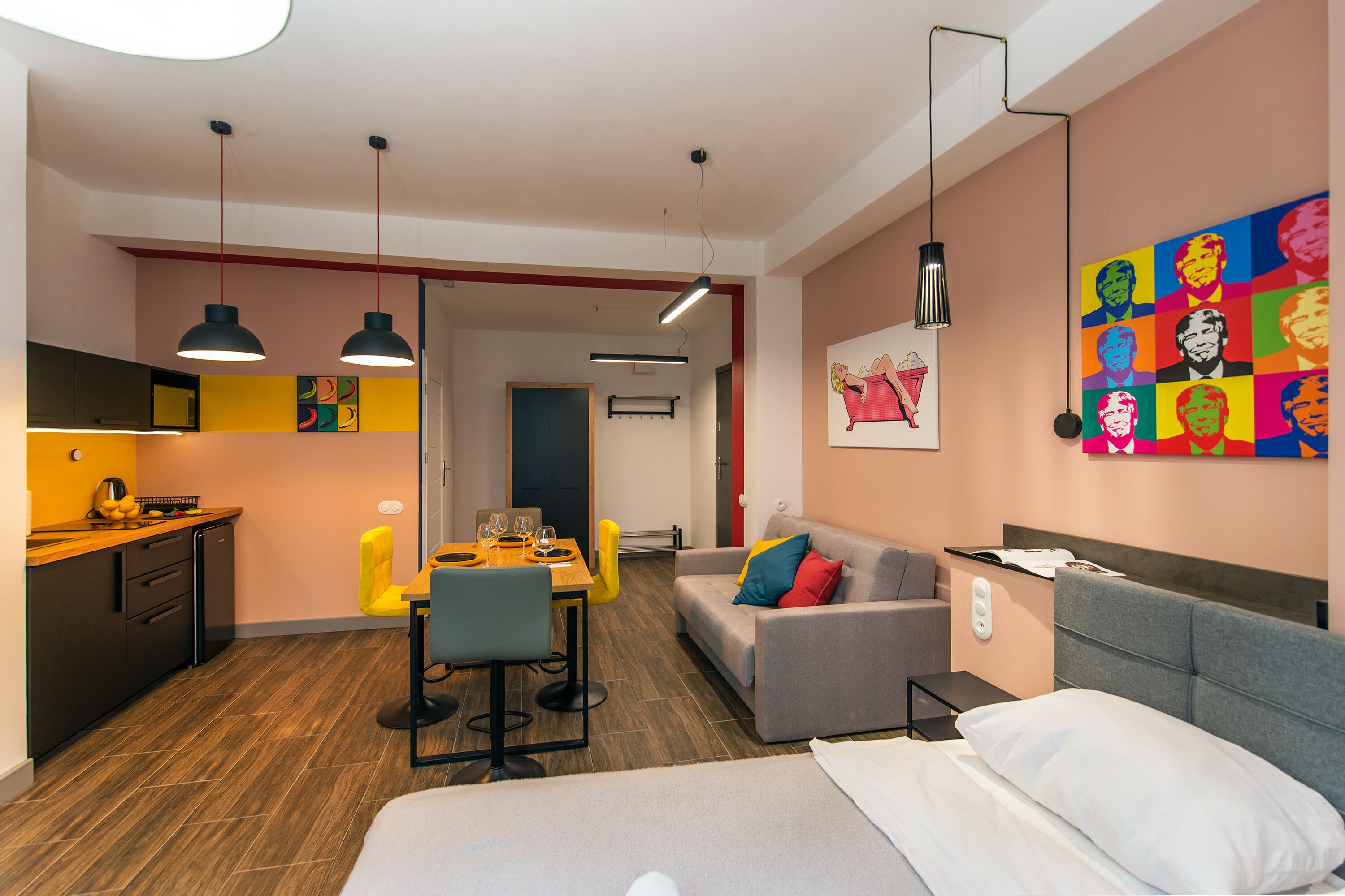 A modern hotel room with a bed, a sofa, a dining area with a table and chairs, a kitchen area, and a colorful artwork on the wall.