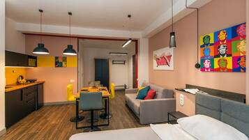 A modern hotel room with a bed, a sofa, a dining area with a table and chairs, a kitchen area, and a colorful artwork on the wall.