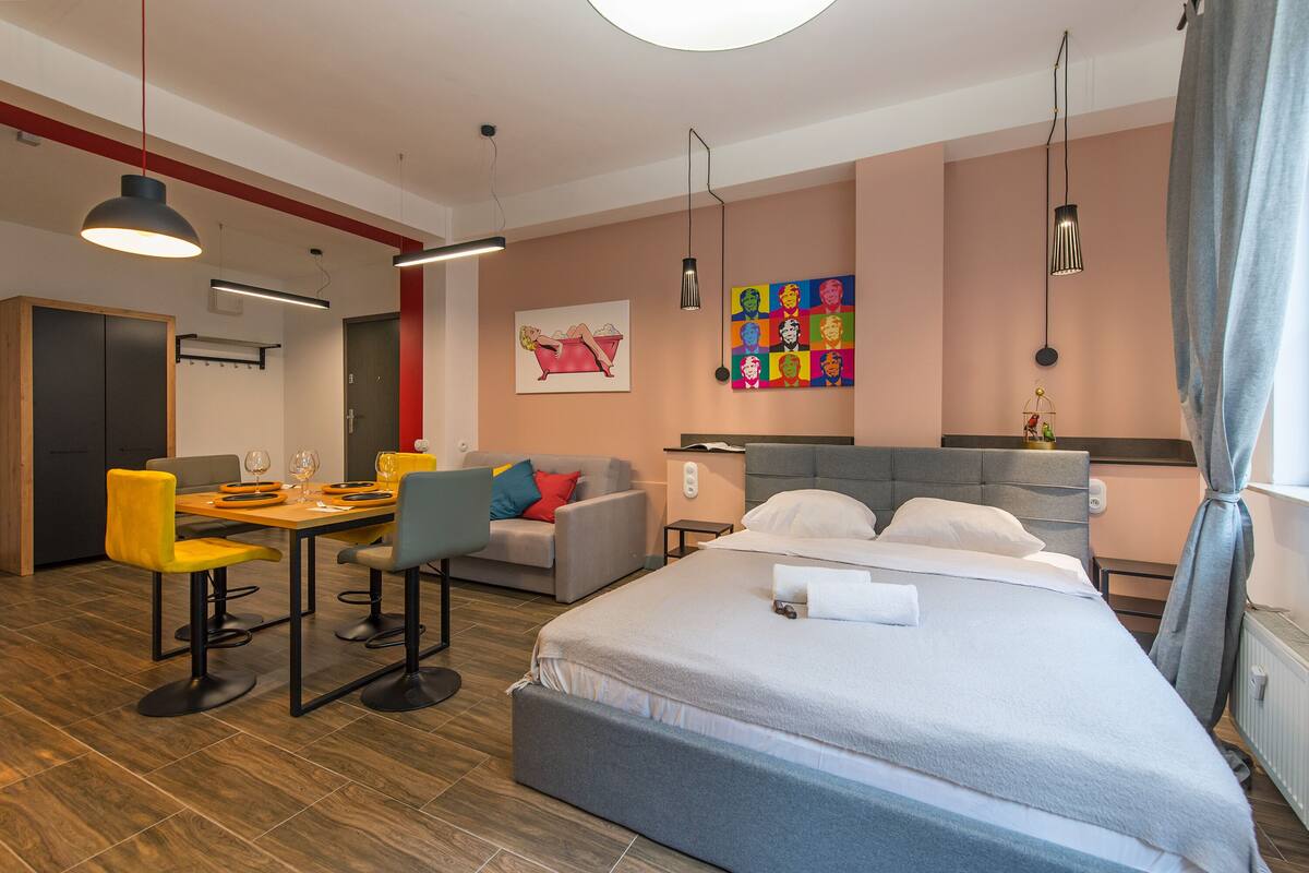 A modern hotel room with a large bed, a dining area with yellow chairs, a gray sofa, and a colorful artwork on the wall.
