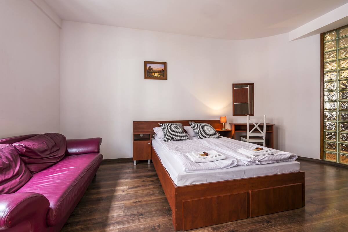 A modern bedroom with a wooden bed, a leather sofa, a nightstand, a bedside table, and a wall-mounted mirror.