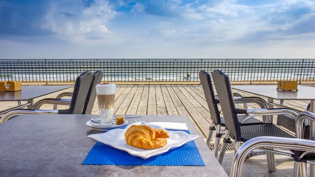 Una mesa con un croissant, una taza de café y una porción de pastel en una terraza con vistas al mar.