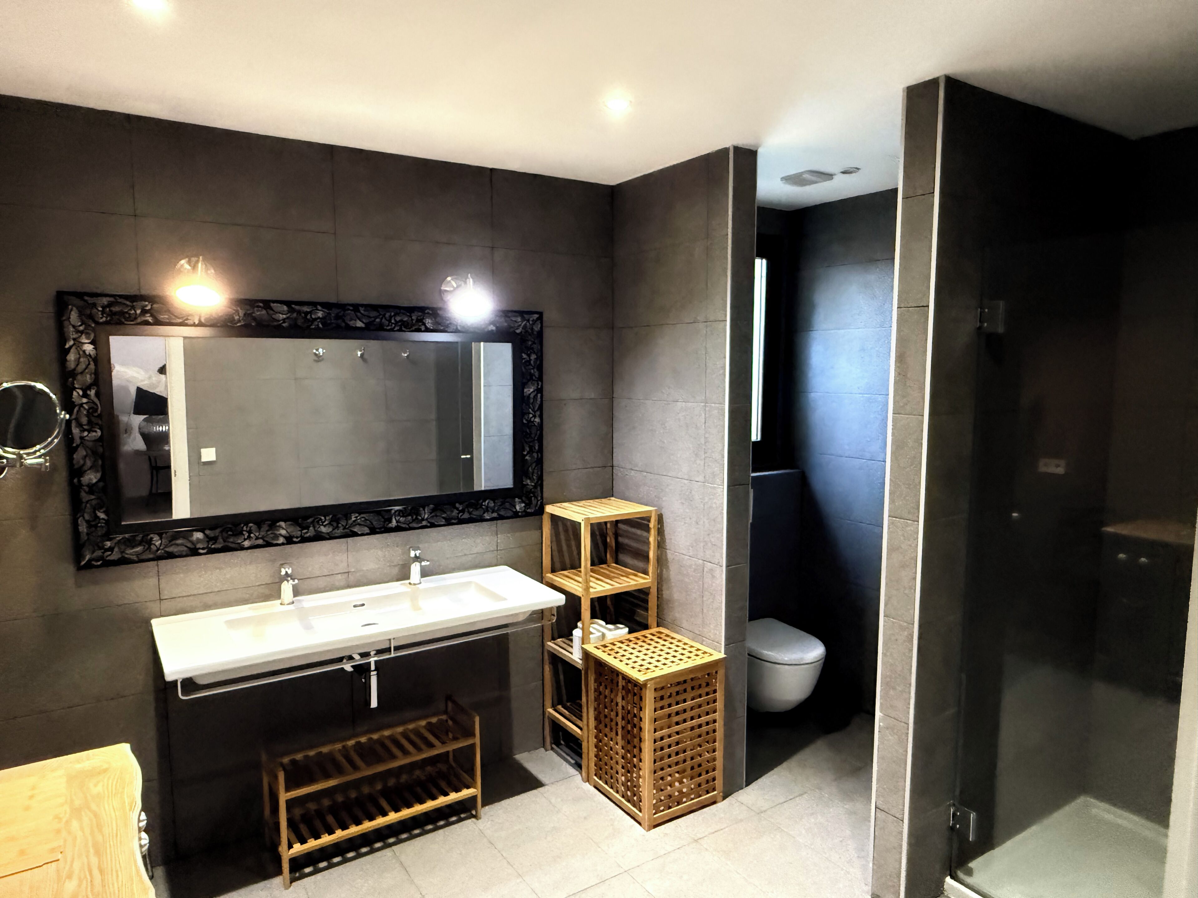 Un baño moderno con un espejo grande, un lavamanos blanco, un taburete de madera y una zona de ducha.