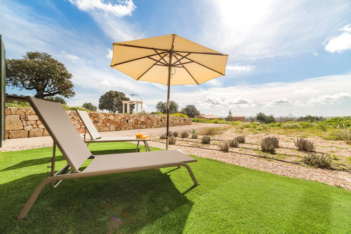 Een terras met een lounger, parasol, een stenen muur en een kleine pergola op de achtergrond.