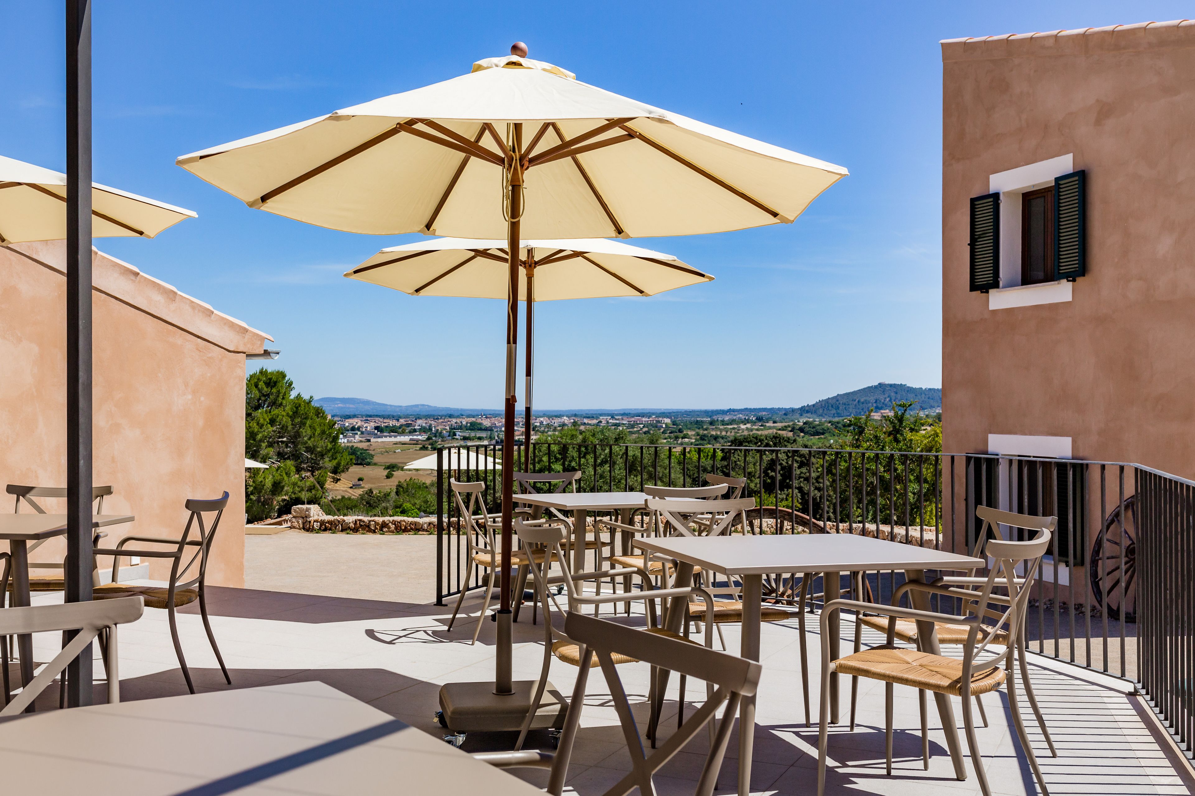 An outdoor patio with tables and chairs, beige umbrellas, and a scenic view of a valley and mountains.