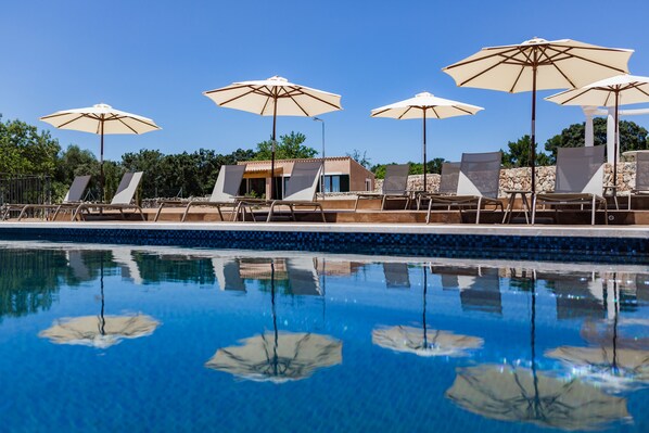 Un espace piscine doté de chaises longues et de parasols, une piscine à l’eau d’un bleu limpide et un mur de pierre en arrière-plan.