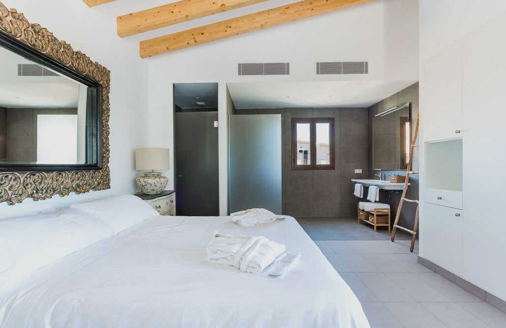 Habitación de hotel con una cama grande, un espejo con marco decorativo, un baño con lavamanos y toallero, y una viga de techo de madera.