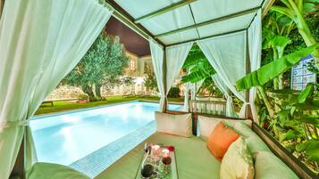 A poolside lounge with a view of a swimming pool, surrounded by lush greenery and a white canopy.