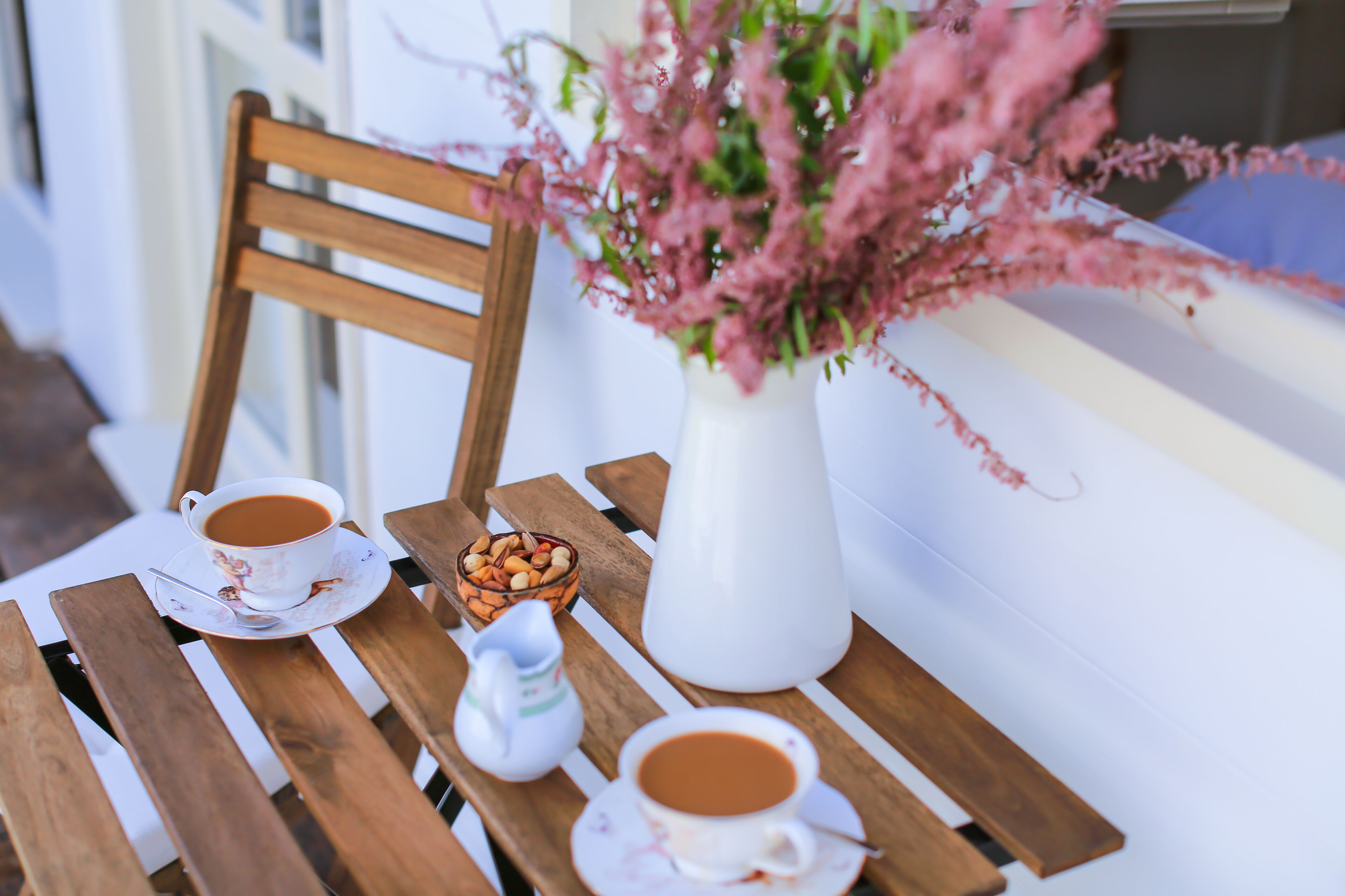 Una mesa de madera con dos tazas de café, un cuenco pequeño de nueces y un florero blanco con flores rosadas.