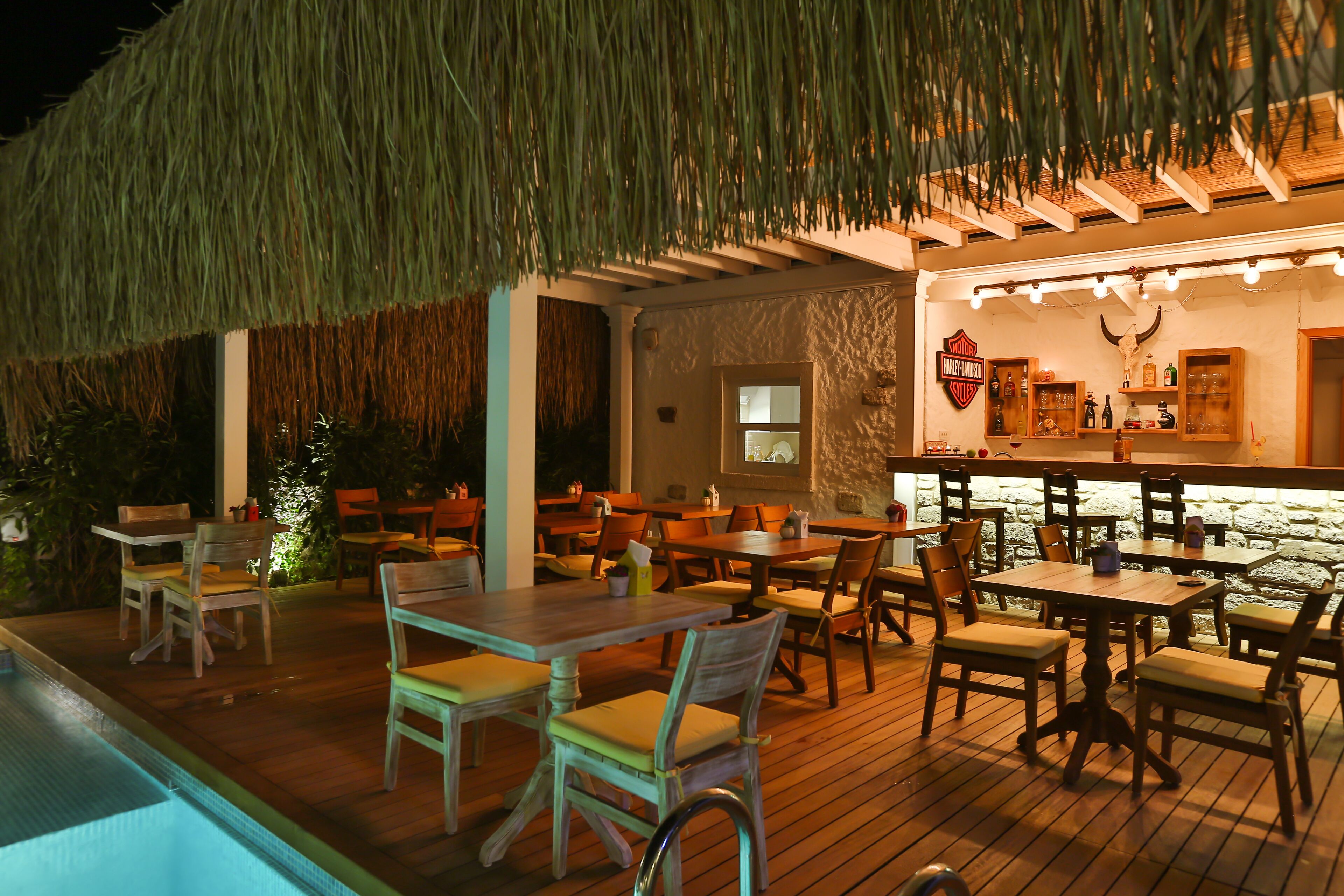An outdoor dining area with wooden tables and chairs, a bar with a stone wall, and a thatched roof.