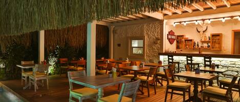 An outdoor dining area with wooden tables and chairs, a bar with a stone wall, and a thatched roof.