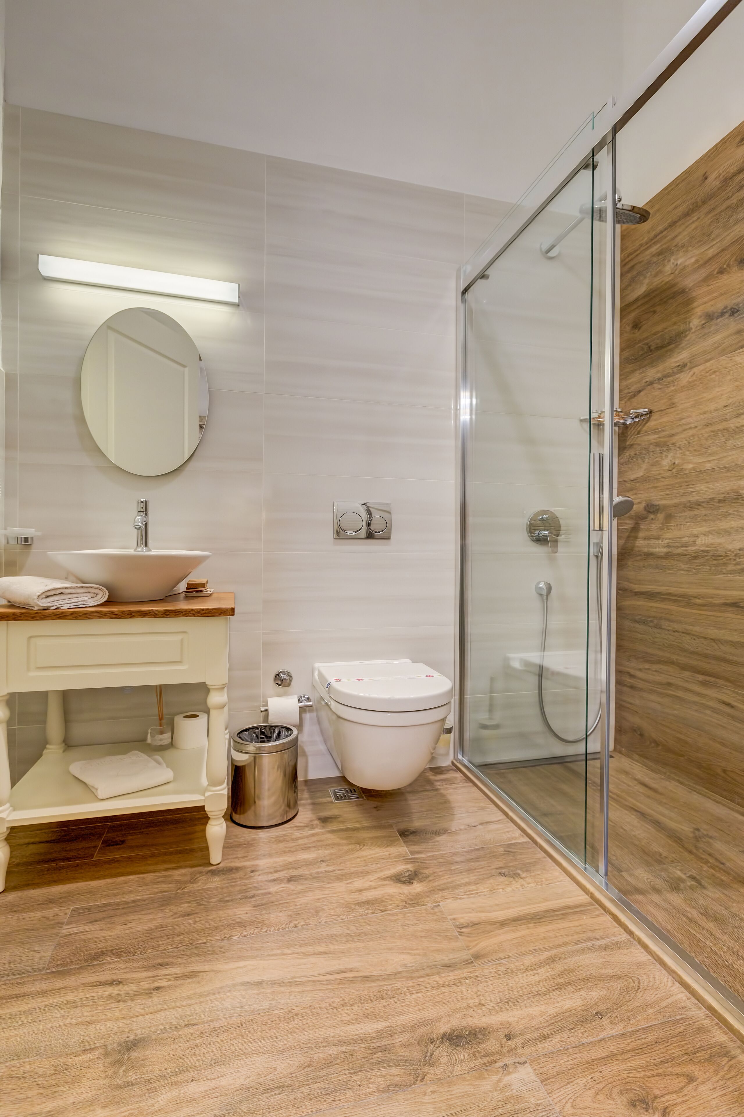 A modern bathroom with a wooden vanity, a round mirror, a white sink, and a glass-enclosed shower.