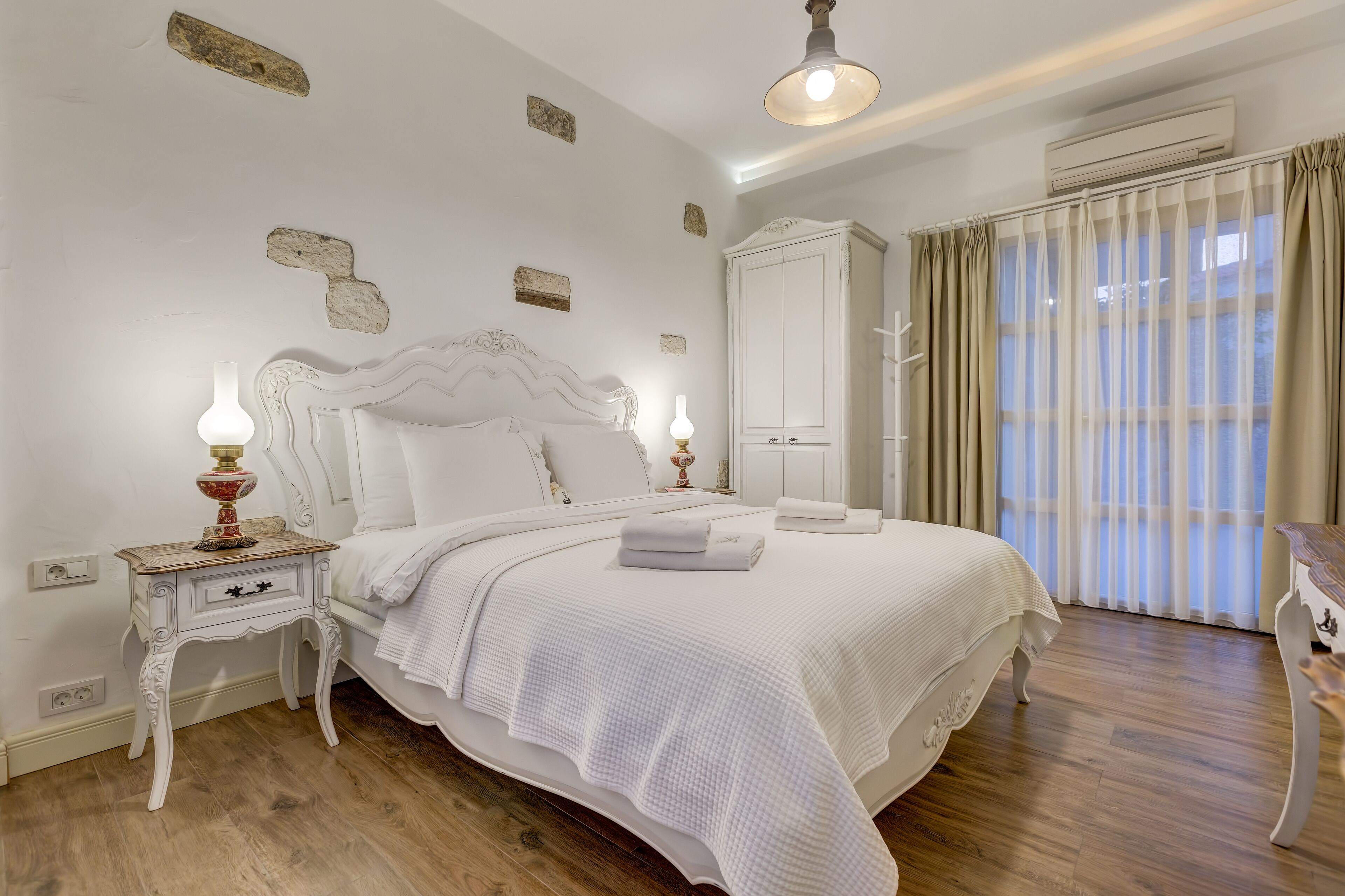 A neatly arranged bedroom with a large bed, a nightstand with a lamp, a wardrobe, and a window with curtains.