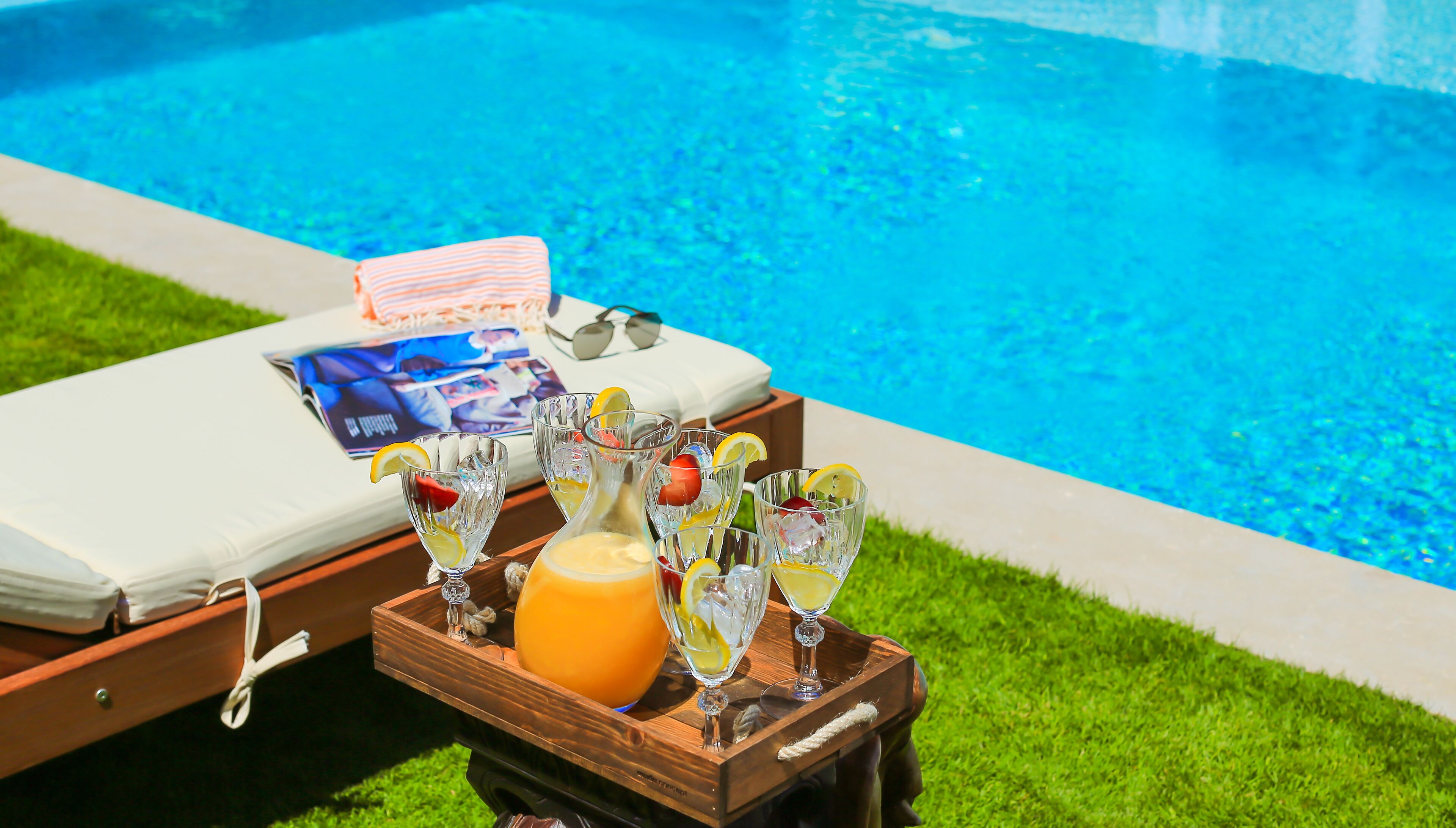A poolside lounge area with a wooden table holding drinks and a magazine, a white lounge chair, and a blue pool in the background.