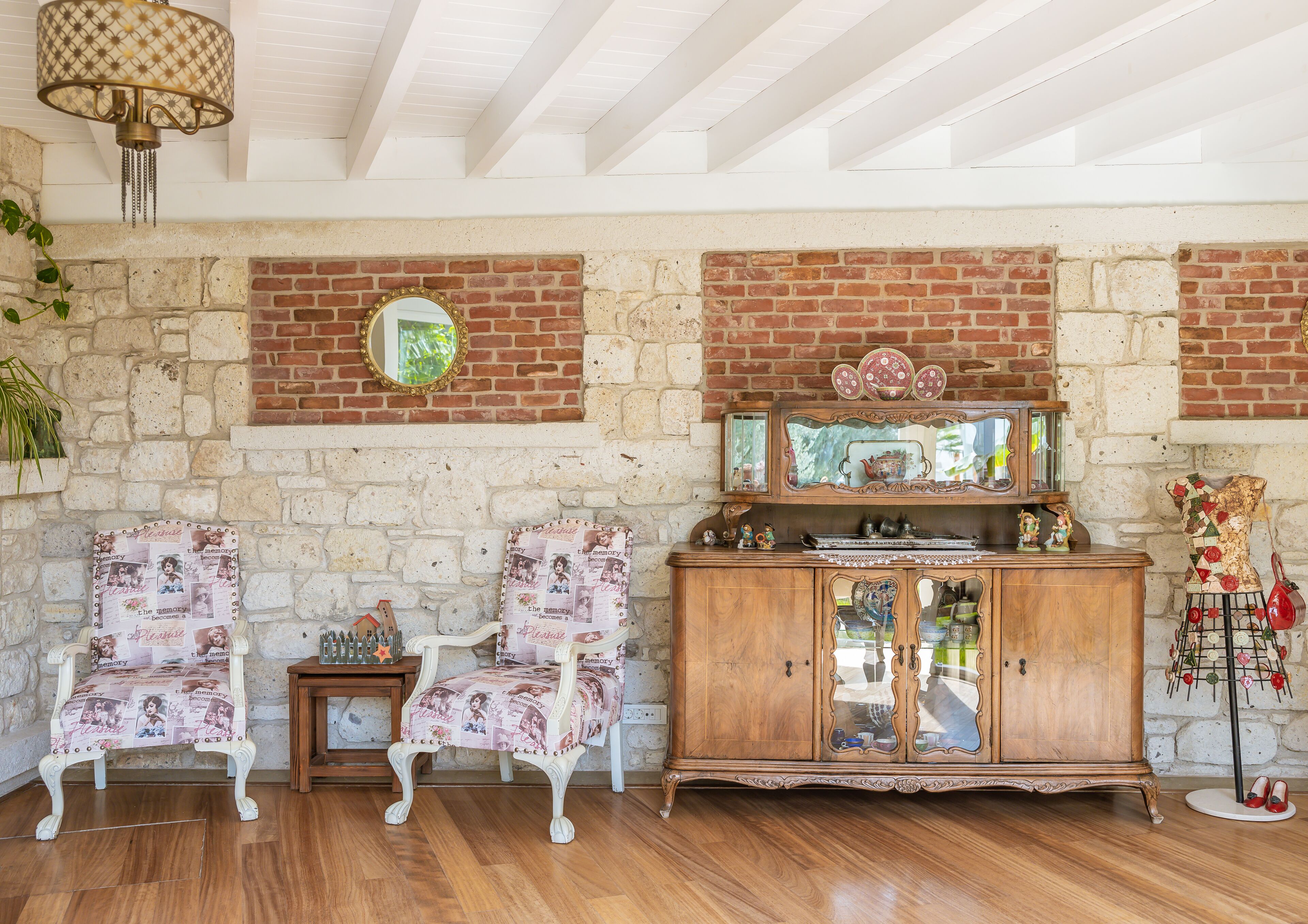 Una habitación con pared de ladrillo, suelo de madera, un mueble de madera con puertas de cristal, dos sillas blancas, una mesita y una lámpara colgante.