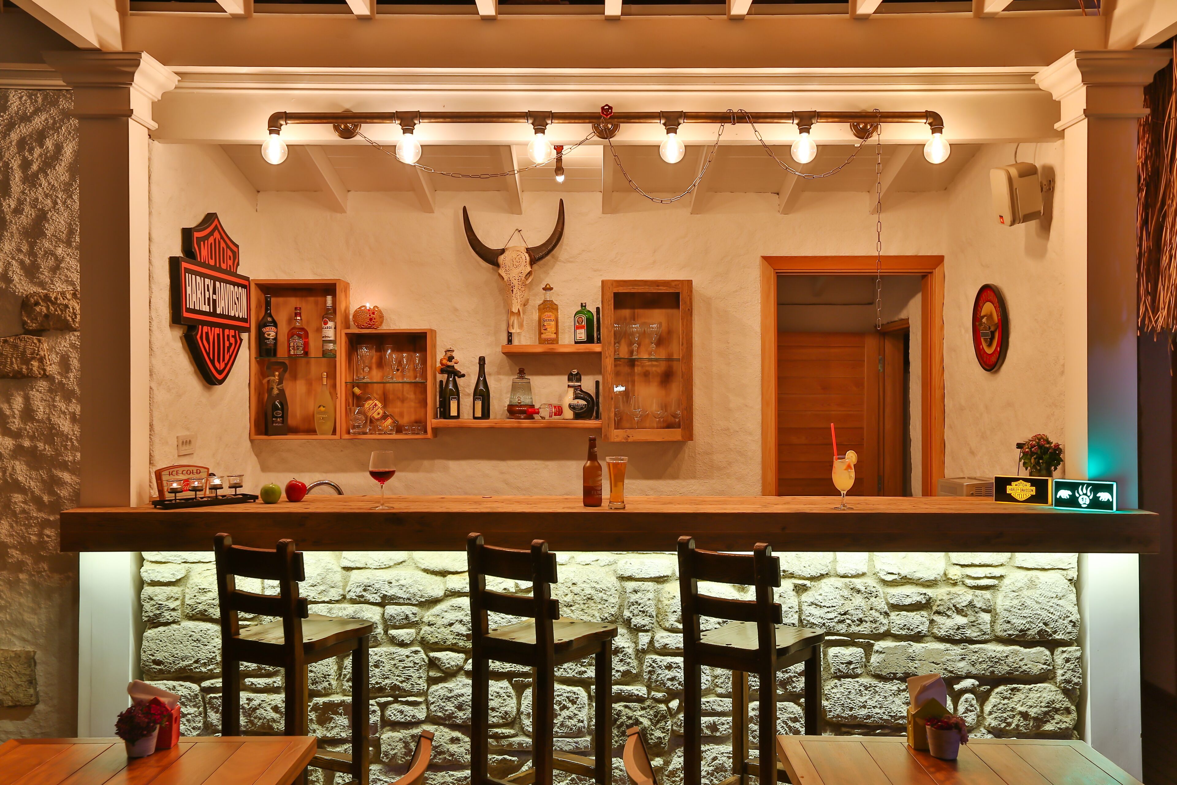 A bar area with a Harley-Davidson sign, wooden shelves with bottles, and a stone wall.