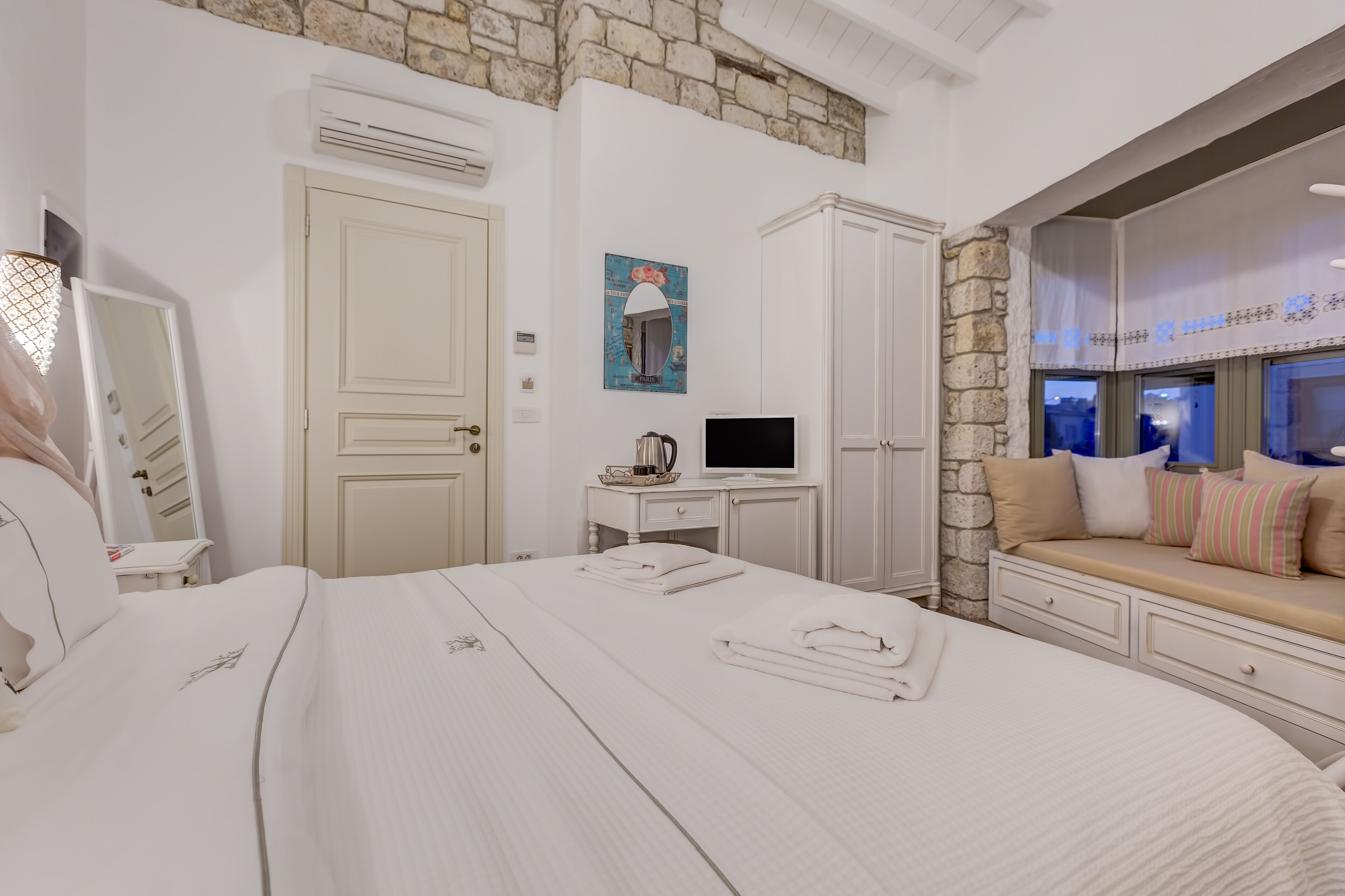A hotel room with a large bed, a desk with a computer, a TV, and a stone wall.