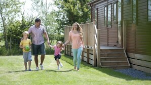 A family walking on a grassy lawn with a wooden deck and a cabin in the background. - Sandy Meadows Lodge Park (Burnham-on-Sea)