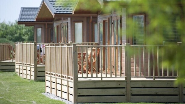 A wooden cabin with a deck and a fenced patio area. - Sandy Meadows Lodge Park (Burnham-on-Sea)