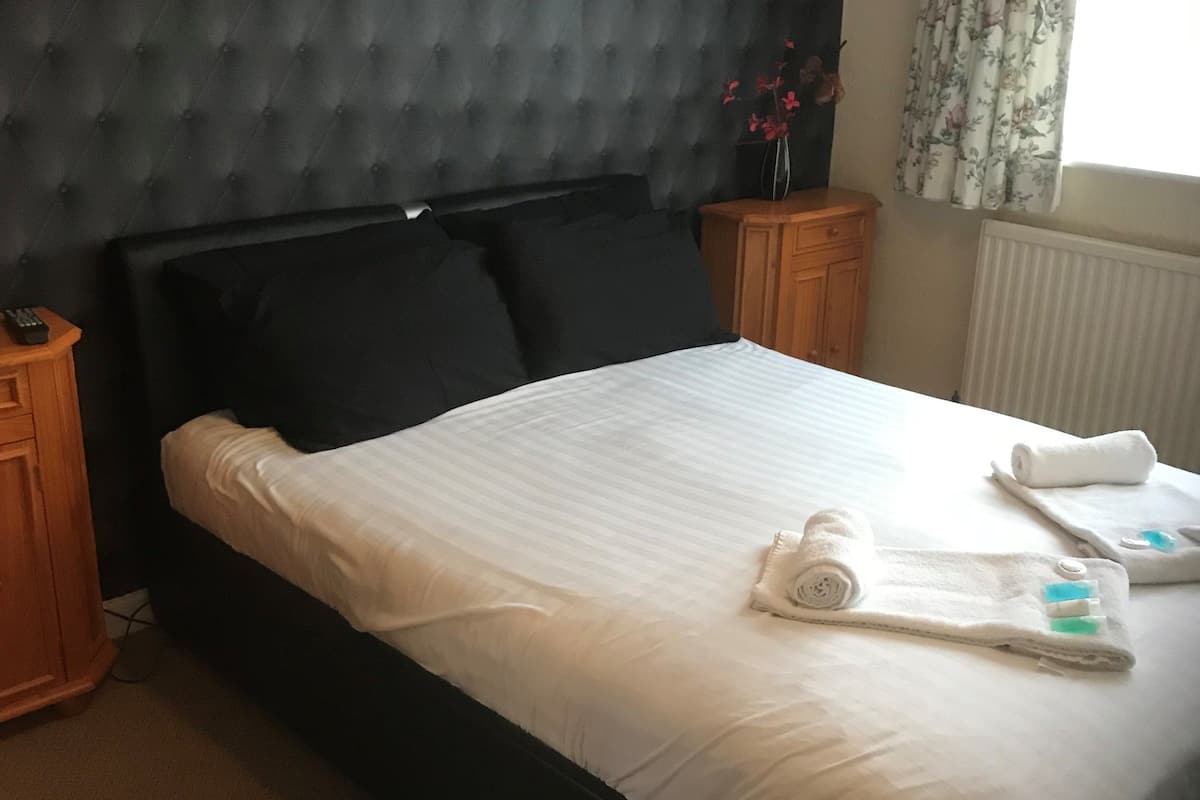 A neatly made bed with white linens and a rolled towel, a tufted headboard, wooden nightstands, and a window with floral curtains.