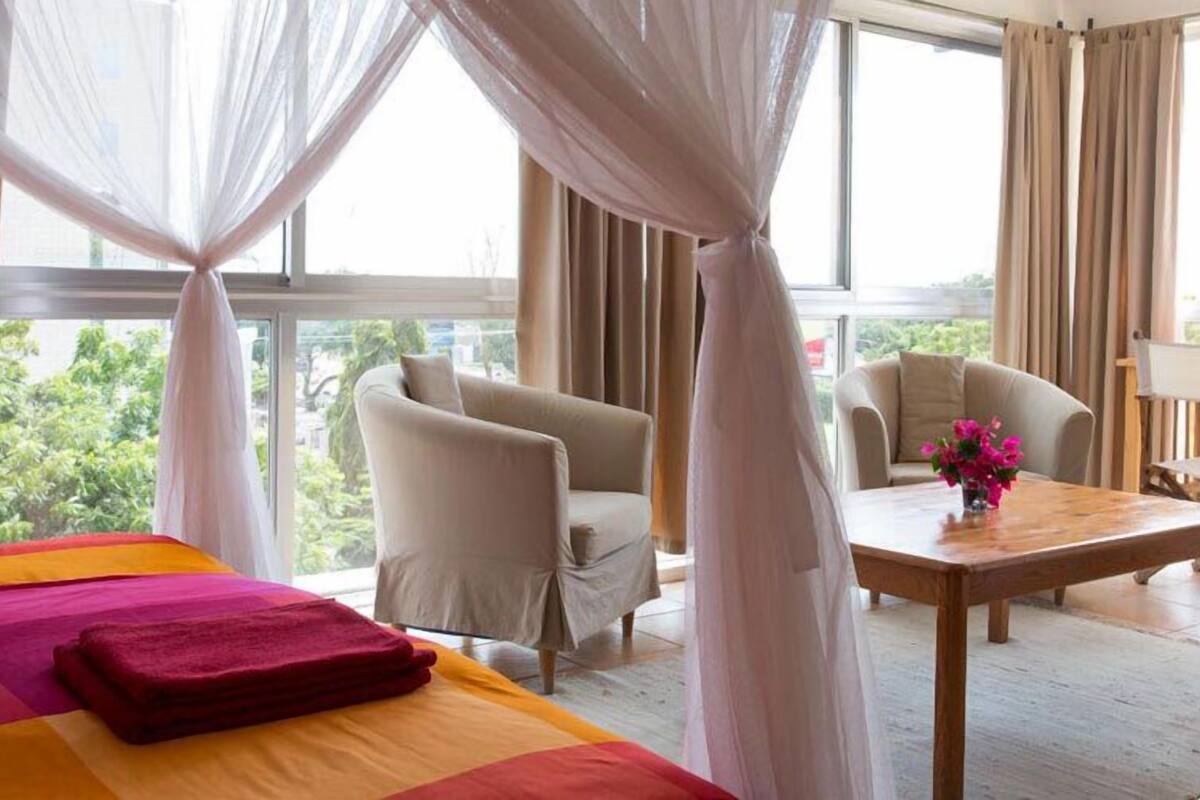 Een hotelkamer met een bed, twee stoelen, een tafel en uitzicht op groen door grote ramen.