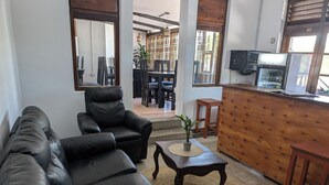 A living room with a leather sofa, a wooden coffee table with a plant, and a kitchen area with a wooden counter and bar stools. - Q-Bar & Guest House (Dar es Salaam)