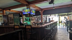 A bar area with wooden flooring, bar stools, and a counter displaying various bottles. There are posters on the wall and a television screen above the bar. - Q-Bar & Guest House (Dar es Salaam)