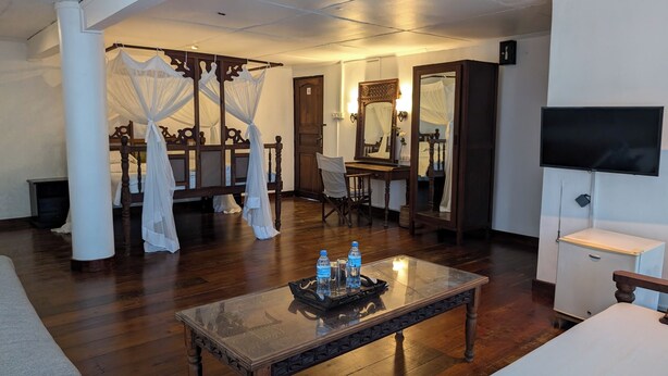 A hotel room with a canopy bed, a sitting area with a glass coffee table, a desk with a chair, a mirror, and a television. - Q-Bar & Guest House (Dar es Salaam)
