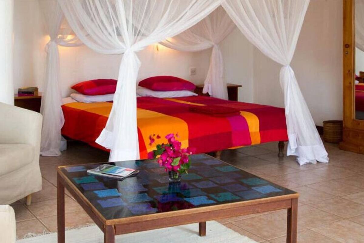 Een kamer met een bed onder een hemelbed, een sofa, een bijzettafeltje met een tijdschrift en bloemen, en een houten nachtkastje.