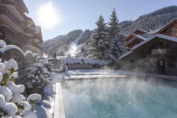 A snow-covered mountain resort with a clear pool area, wooden cabins, and a view of the snowy landscape. - Hôtel L'Eterlou (Les Allues)
