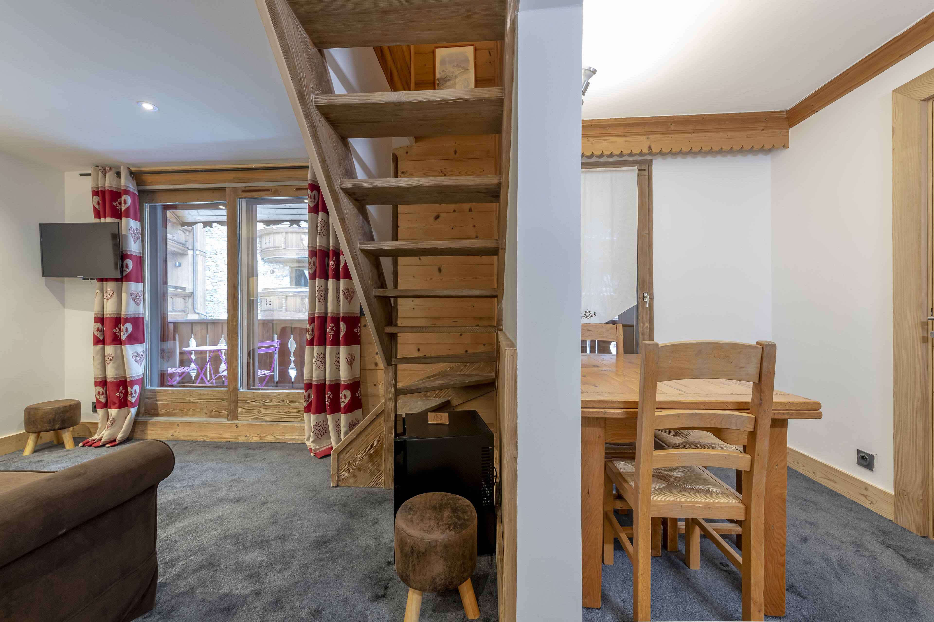 Una escalera de madera que lleva a un balcón con cortinas rojas y blancas, una mesa de comedor de madera con sillas y un pequeño otoman.