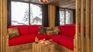 Interior acogedor de una cabaña de madera con un sofá seccional rojo, una mesa de centro de madera con aperitivos y vistas a un paisaje nevado a través de amplias ventanas.
