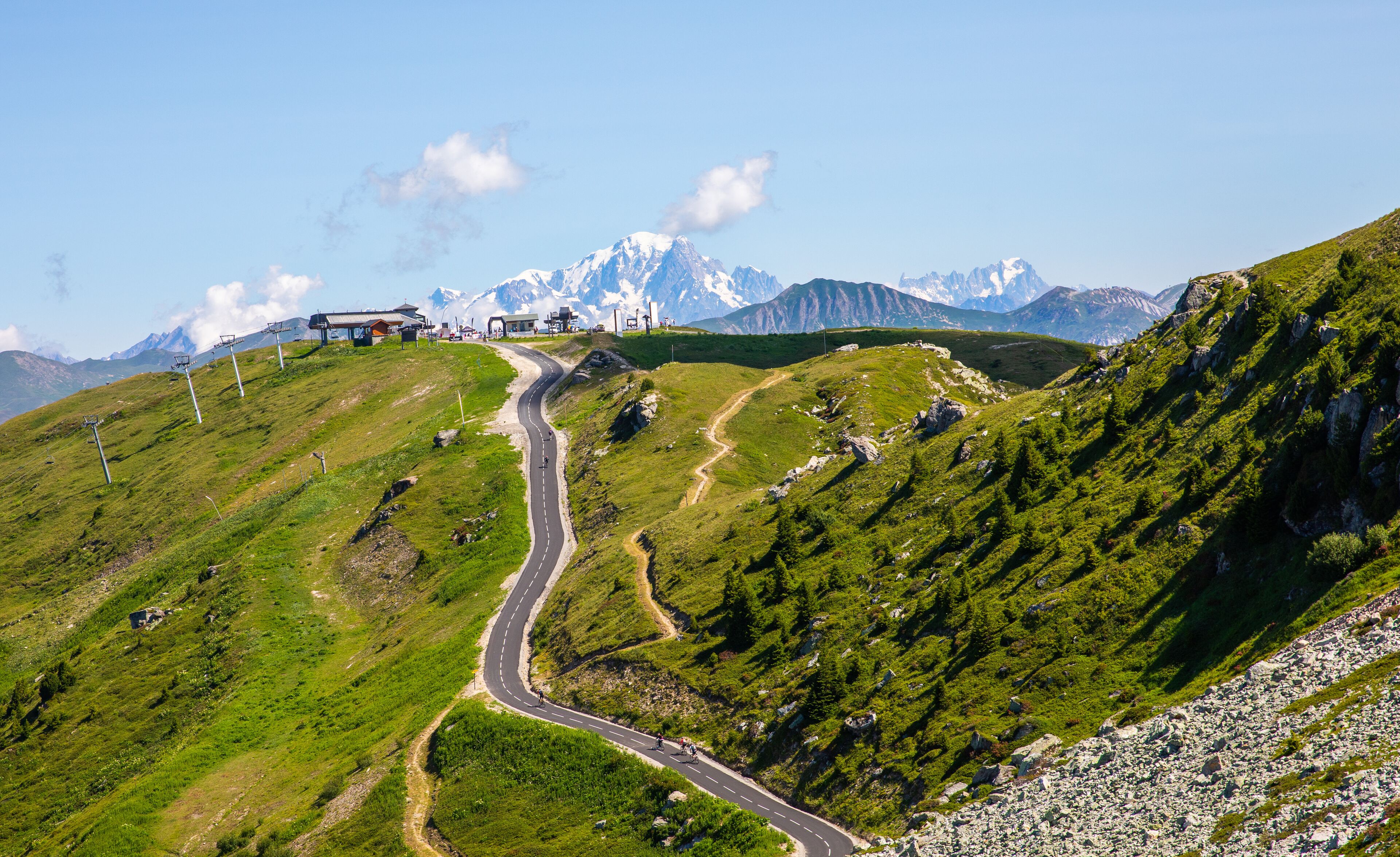 Une route de montagne sinueuse, avec un petit bâtiment perché sur une colline, entouré de collines verdoyantes et de montagnes enneigées en arrière-plan.