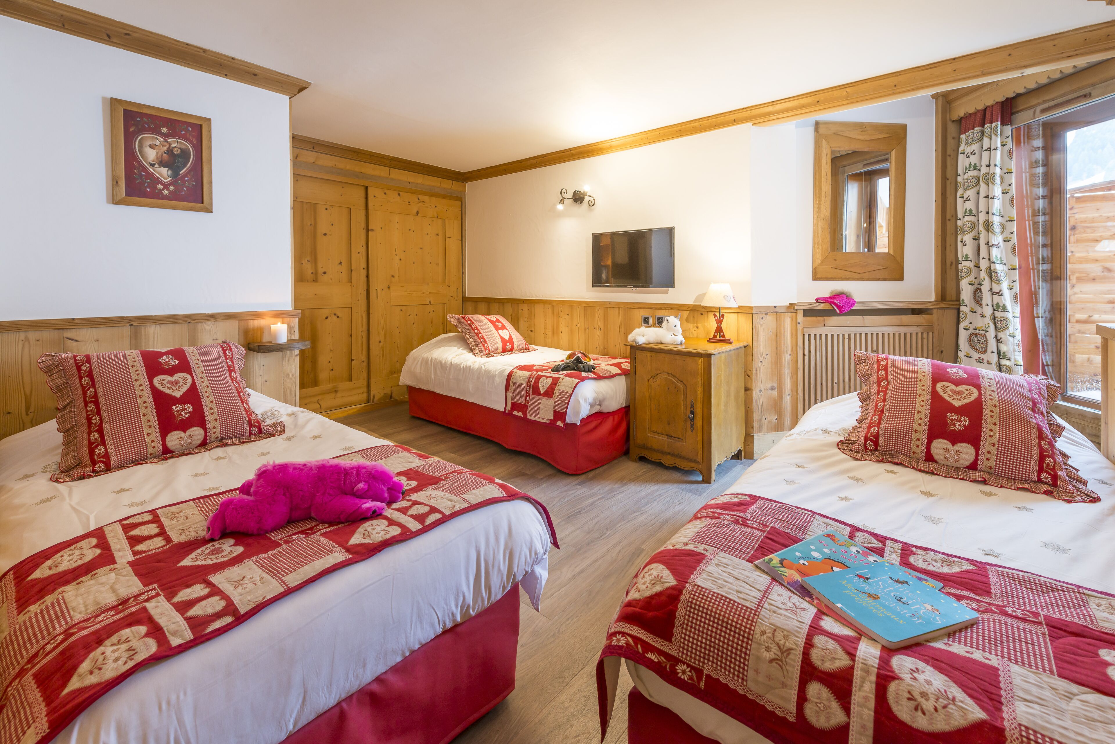 Une chambre accueillante avec deux lits, chacun recouvert d’une couette rouge et blanche, un ours en peluche rose sur l’un des lits et un livre sur l’autre.