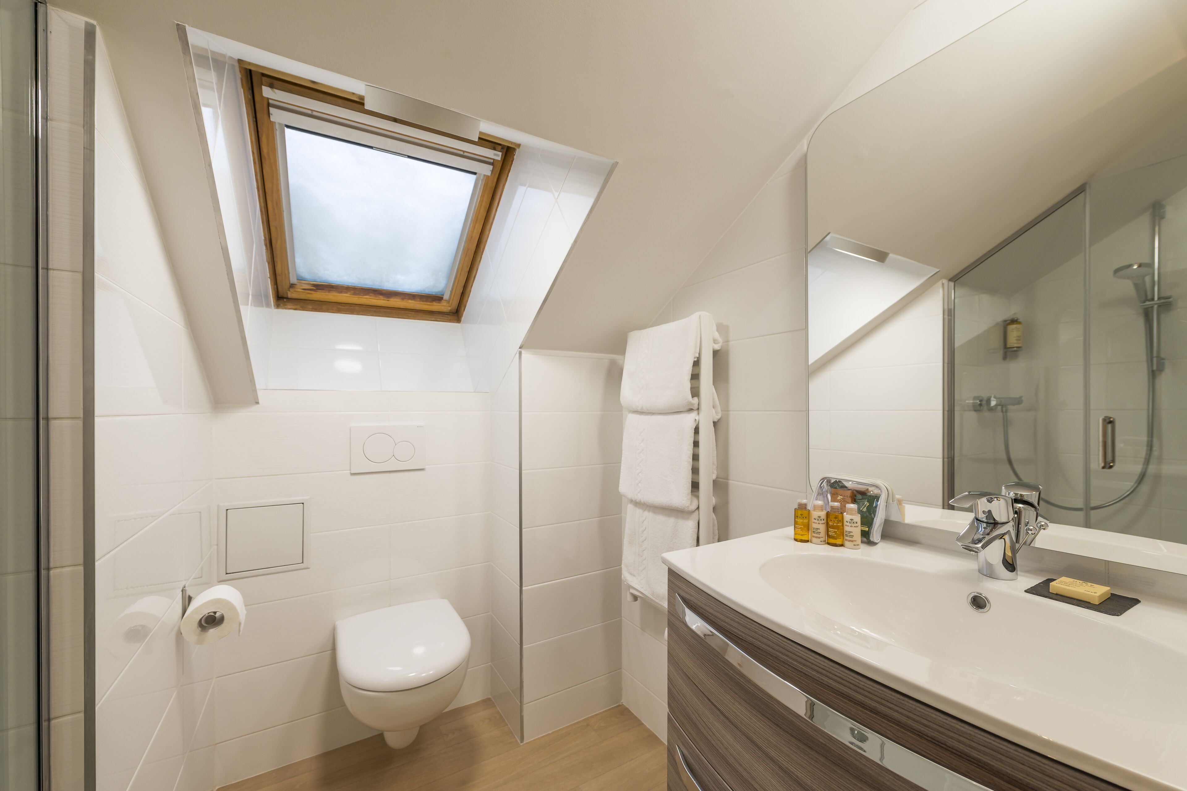 Een moderne badkamer met dakraam, witte tegels, toilet, wastafel met toiletartikelen en een douchecabine van glas.