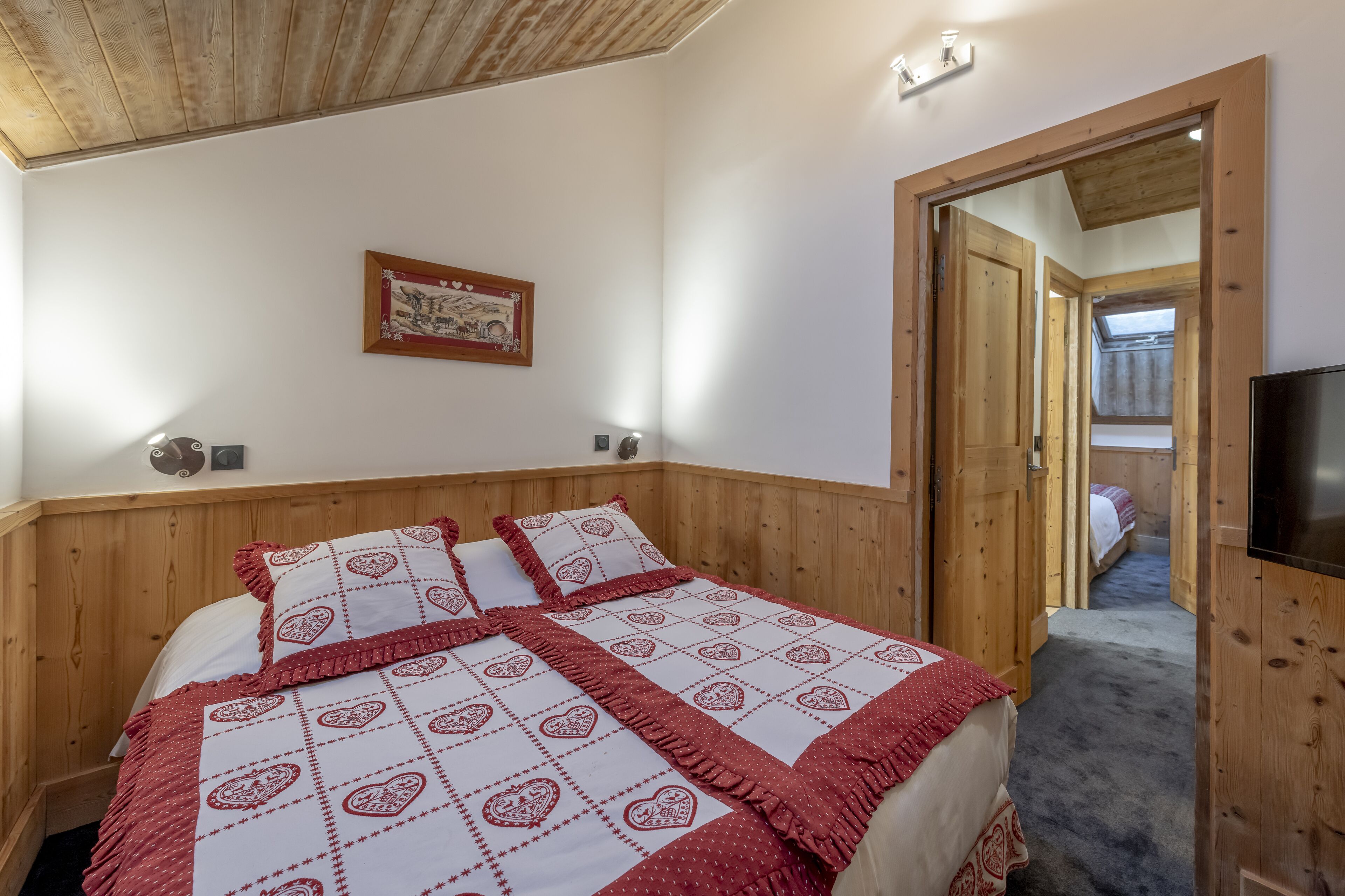 A bedroom with a wooden headboard, a quilted bedspread, a framed picture on the wall, and a television on the right.