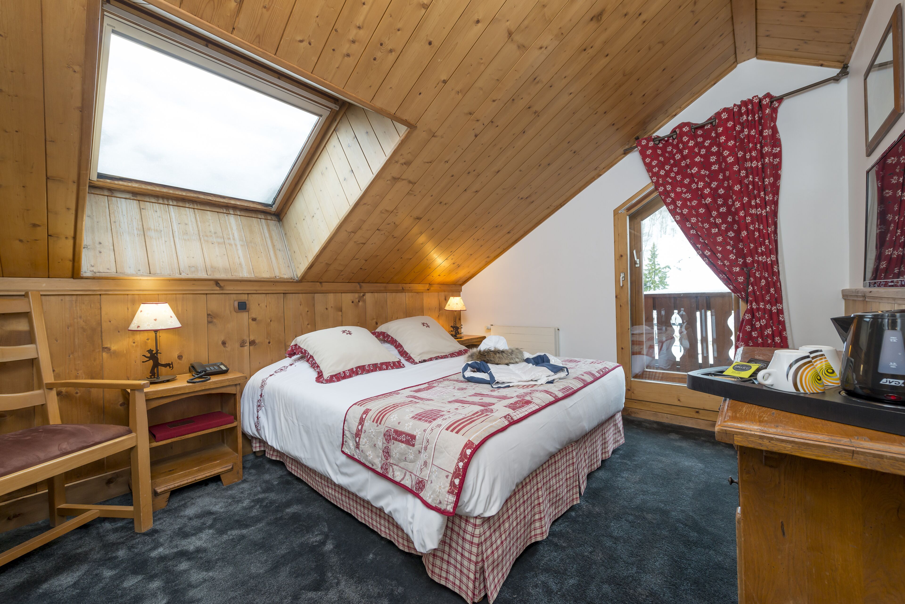 A cozy bedroom with a sloped ceiling, a large window, a bed with a red and white quilt, a bedside table with a lamp, and a view of the outdoors.