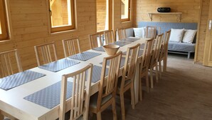Una cabaña de madera con una mesa de comedor dispuesta para seis personas, rodeada de sillas. Se observa un sofá al fondo y una pequeña estantería con objetos.