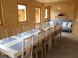 A wooden cabin with a dining table set for six, surrounded by chairs. There is a sofa in the background and a small shelf with items on it.