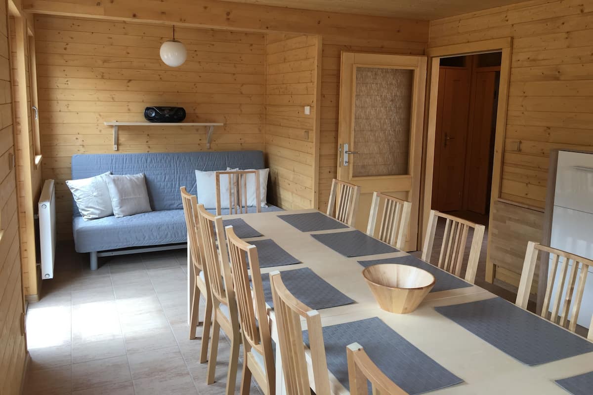 Interior de una cabaña de madera con una mesa de comedor, sillas, un sofá y un ventanal grande.