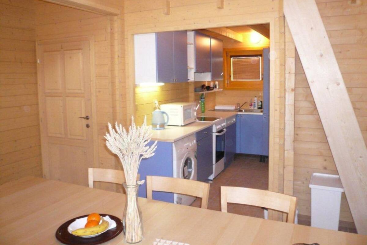 Un interior de madera con una mesa de comedor, sillas, un florero con plantas secas y una zona de cocina con armarios, microondas y lavadora.