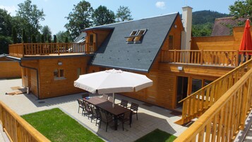 A two-story wooden house with a balcony, a dining area with a table and chairs, and a covered patio.