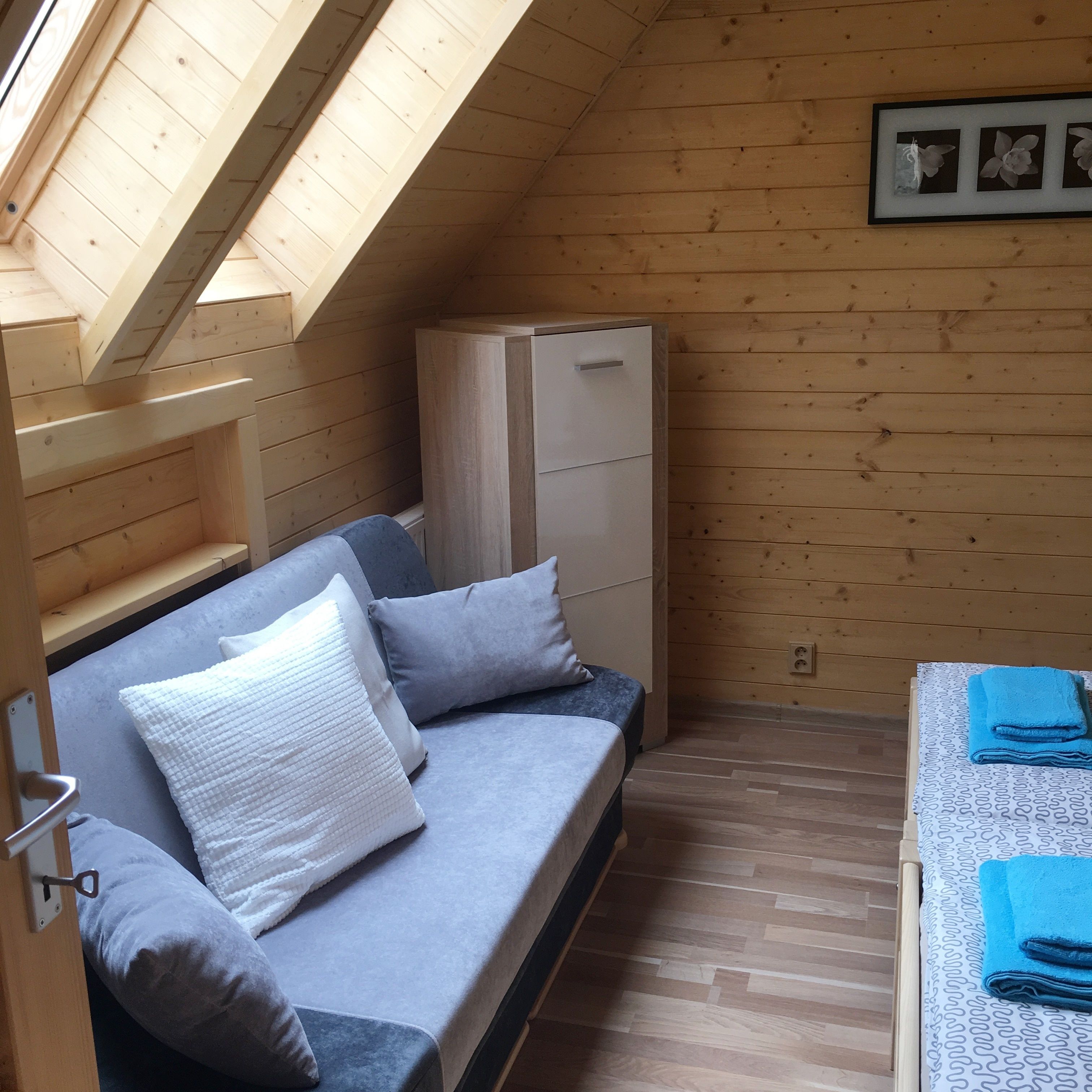 Una habitación compacta con techo inclinado, una cama con ropa de cama gris, un pequeño armario blanco y un cuadro en la pared.
