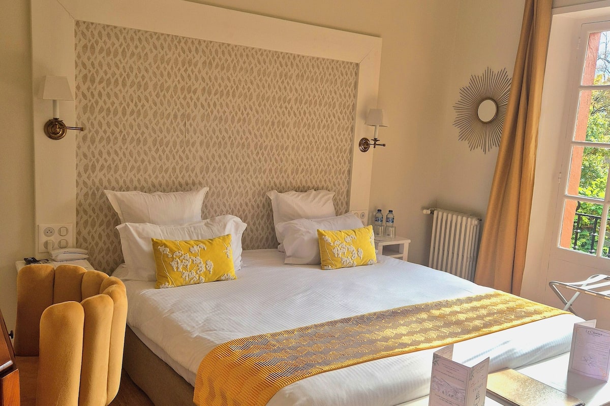 Habitación de hotel con una cama grande, dos mesitas de noche, una silla, un radiador y una ventana con cortinas.